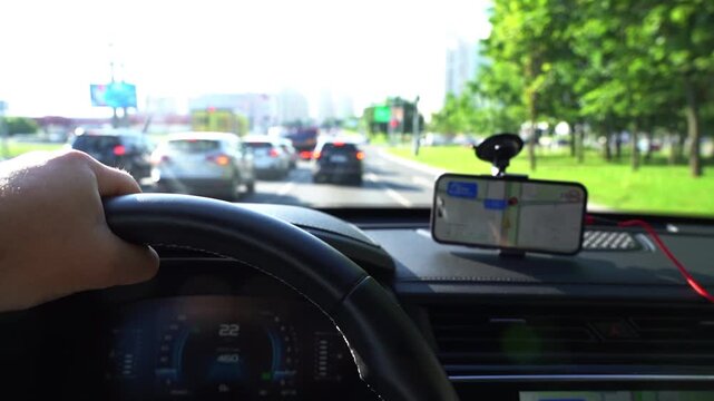 Driver grips steering wheel, focused on road ahead. Phone mounted, displaying navigation map. Cars move slowly through urban traffic. Scene suggests commuting or navigation during rush hour