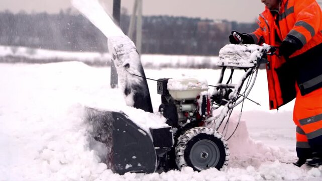 Snowblower operator clears snow-covered path. Person wears bright orange safety gear. Snow flurries around machine and boots. Cold winter day outdoors