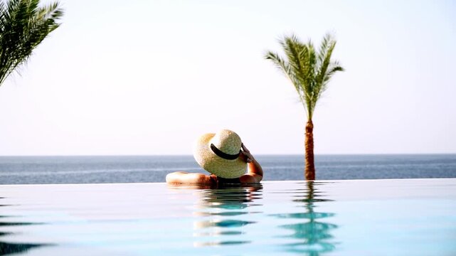 Woman relaxes in infinity pool beside ocean. Palm trees frame tranquil seaside scene. Soft light enhances peaceful vacation atmosphere. Ideal for luxury travel or wellness campaigns
