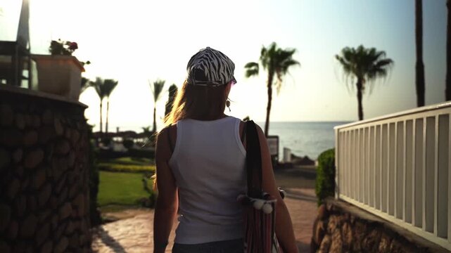 Young woman walks toward ocean with striped cap. Sunset glows behind palm trees and stone walls. She carries bag, wears white tank top, denim shorts. Solemn pace suggests travel, quiet reflection