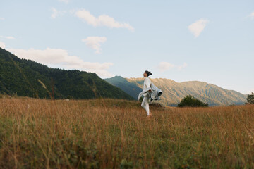 Obraz premium Woman walking in a grassy field near mountains under a blue sky with clouds. Lifestyle concept with nature, freedom, and outdoor activity in soft natural light.