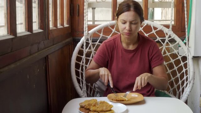 A calm woman cuts a breaded cutlet at a white table. She feels content and relaxed sitting inside a cozy hanging swing chair. The wooden porch feels peaceful and quiet.