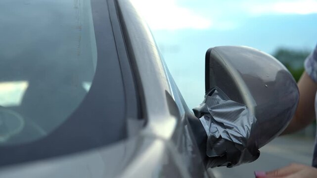 Desperate woman hands wrapping grey duct tape around broken car side mirror after accident, temporary and unsafe roadside repair before heading to auto repair shop, close-up, slow motion.