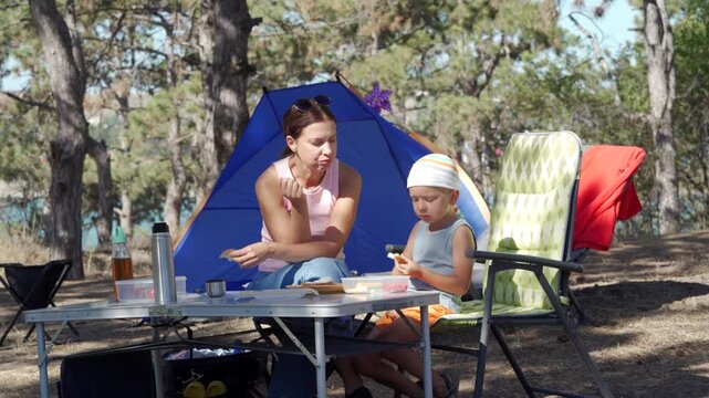 A relaxed mother and her son happily eat bread at a picnic table in a sunny forest campsite. They feel content and peaceful sharing a cozy outdoor breakfast next to their blue tent.