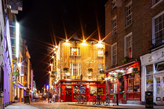 Night view of the iconic red-fronted "The Temple Bar" pub in Dublin, its upper facade covered in warm fairy lights, with a Guinness advertisement, cobblestone street, parked bicycles