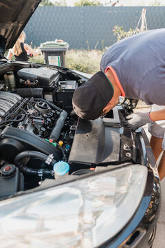 Mechanic leaning over open hood inspecting engine compartment of parked sedan in residential driveway under daylight, checking coolant reservoir, air intake, wiring, and front components