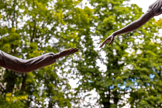 Bronze Sculpted Hands Reaching Toward Each Other. Two bronze sculpted arms reach toward one another against a softly blurred canopy of sunlit green trees.
