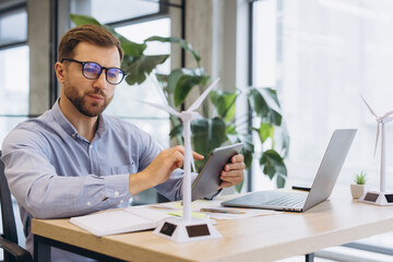 Obraz premium Male engineer using a tablet, working on renewable energy project with wind turbine models in a modern office
