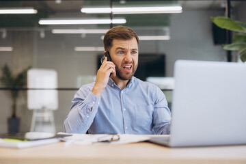 Fototapeta premium Stressed businessman talking on smartphone, reacting with anger and frustration to bad news, working on laptop in modern office