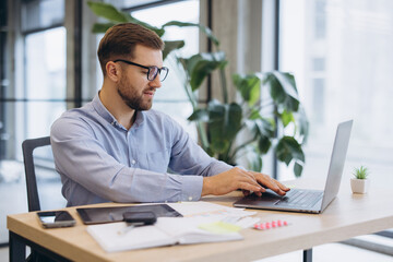 Fototapeta premium Young businessman working at desk, using laptop in modern office, focusing on productivity and technology