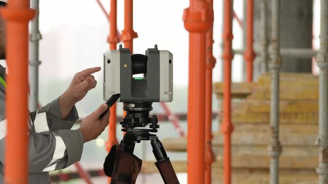Worker points to surveying instrument on tripod. Construction site features orange scaffolding and blurred structures. Professional focus during field measurement work