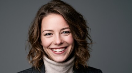 Naklejka premium Portrait of a smiling young woman with wavy brown hair wearing a turtleneck sweater against a grey background