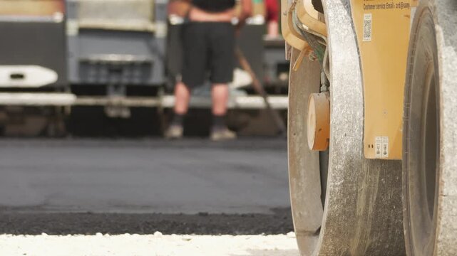 Heavy road roller compacts fresh asphalt. Worker focuses on the road surface. The truck's rear is visible in the background. Smooth black asphalt spreads evenly. Ideal for construction projects