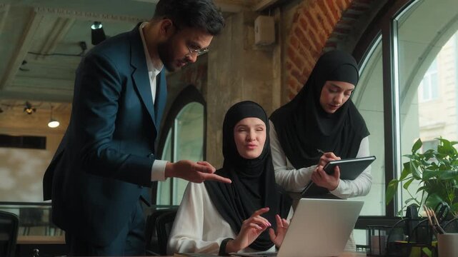 Hindu business team in office male boss mentor talking to intern female pointing in laptop explain businessman with businesswomen in hijab advice with computer project corporate teamwork cooperation
