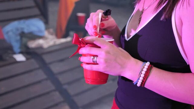 Close-up of a woman's hands holding a red plastic cup with a bow and a straw at an outdoor party.