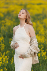 Obraz premium Pregnant woman in flowing white dress and knitted cardigan stands in yellow flower meadow with eyes closed and hands on belly, showing calm anticipation, gentle natural light, blurred spring