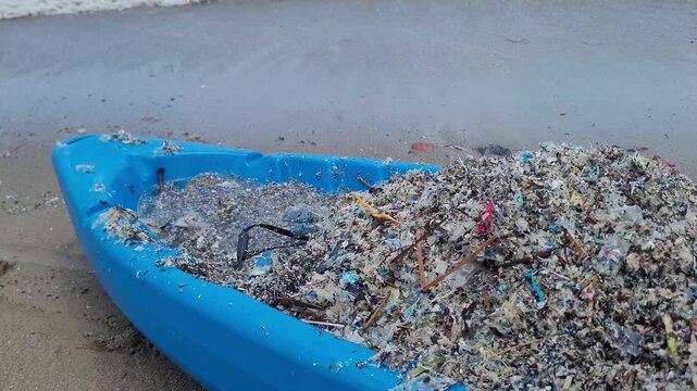 A blue boat filled with collected microplastics and marine debris sitting on a sandy beach near the sea with waves.