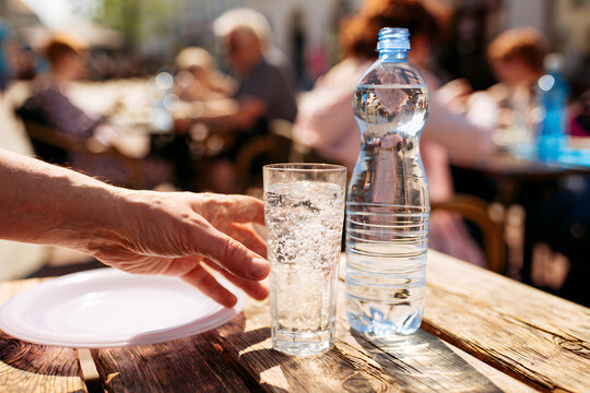 Someone stretches out to grab the water bottle from the cafe table. The glass swells with bubbles. Diners enjoy their meal against the indistinct outdoor restaurant backdrop. A sunny day with refreshi