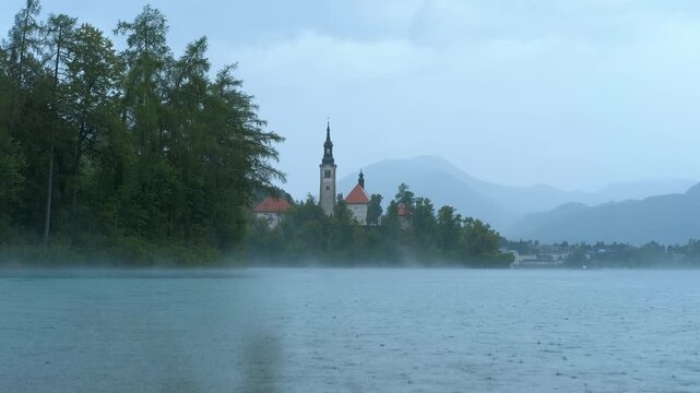 Rain falling on lake bled island in slovenia. Gentle rain creates a mystical atmosphere over lake bled island, blurring the picturesque church and surrounding trees against a backdrop of mountains