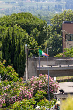 Italian flag waving in wind surrounded by green cypress trees and blooming pink flowers near concrete bridge during sunny summer day in Italy landscape.