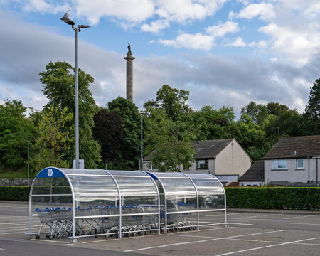 Duke of Gordon monument standing tall over the Tesco Supermarket parking lot in Elgin under a bright daytime sky with lush green trees and clouds