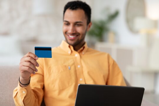 Smiling Young Man Holding Credit Card While Shopping Online With Laptop At Home. Engaged In Online Payment Process At Cozy Living Room, Demonstrating Modern Shopping Behavior, Business Concept.