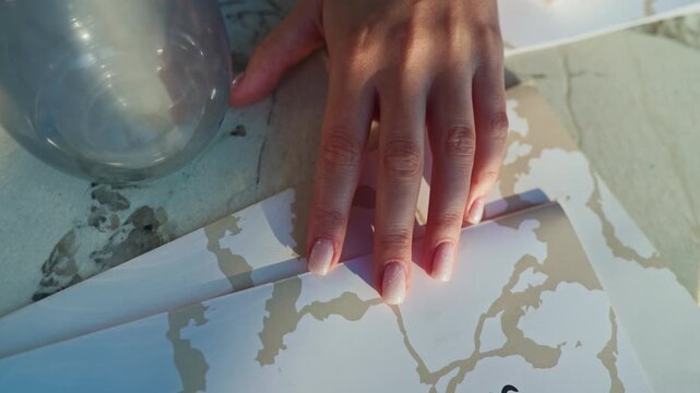 Hand with pink manicured nails resting on printed map papers beside glass bowl in soft sunlight