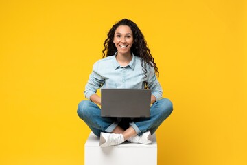 Fototapeta premium Smiling Young Mixed Race Woman Sitting Cross-legged On Cube While Working On Laptop In Studio With Bright Yellow Background. Joyful Emotion Conveyed In A Modern Remote Work Concept.