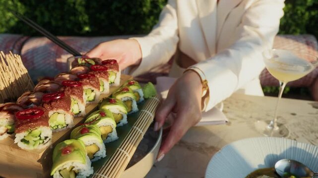 Outdoor sushi platter with avocado rolls, elegant hand serving food beside drink on sunlit terrace table