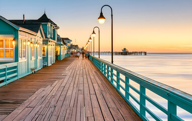 Obraz premium Oceanfront boardwalk with light blue buildings, shops and lamp posts at sunset. People walk along the wooden pier near the sea. Relaxing coastal vacation scene