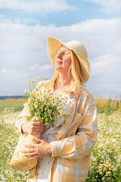 Blonde woman in straw hat, plaid shirt, and floral dress stands with her eyes closed in chamomile field, clutching bouquet of daisies and wicker bag, against blue sky with clouds