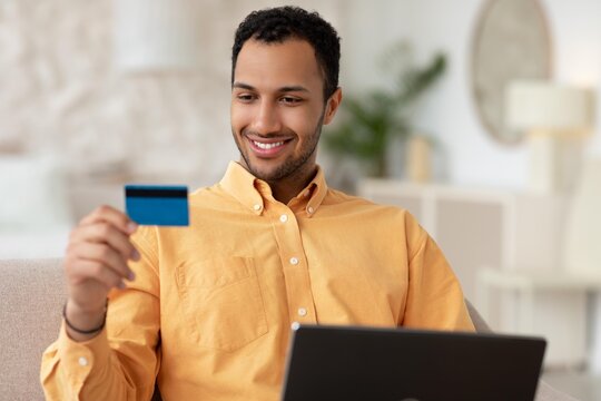 Smiling Young Black Man Holding Credit Card While Looking At Laptop In Cozy Home Office Setting. Engaged In Online Shopping Or Payment Process In Modern Indoor Workspace. Business Concept.