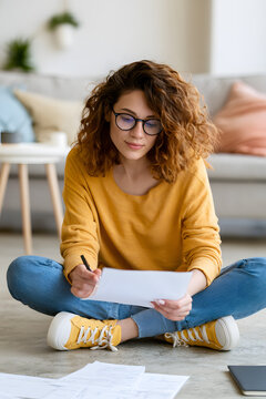 A vertical image of a young adult Caucasian woman seated on the floor, assessing her bills and notes as she manages household finances and copes with financial stress. Perfect for promoting informatio