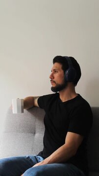 Vertical detailed view of a latin man sitting on a grey couch using headset and drinking something on a white cup in a peaceful attitude