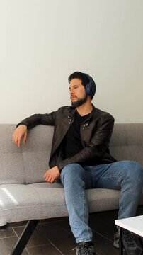 Vertical view of a man wearing a leather jacket sitting on a grey couch listening music on a wireless headset in a serious attitude