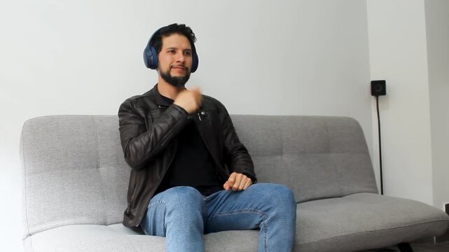 A hispanic man sitting on a grey couch listening music on a headset and doing drums air in a happy attitude