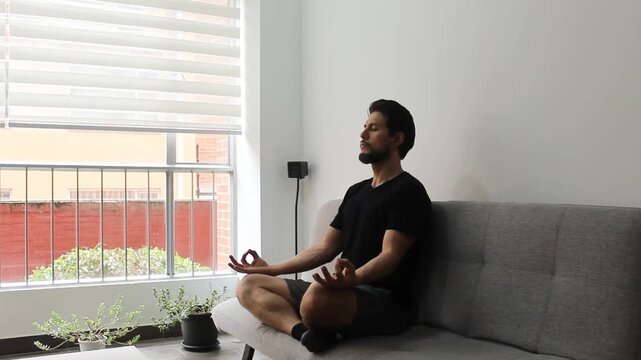 A latin man sitting on a couch in the lotus pose meditating in the morning
