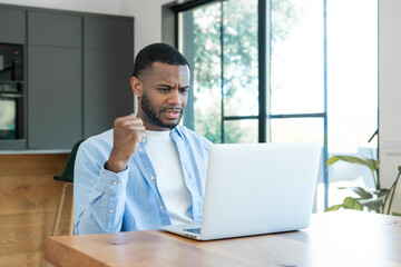Fototapeta premium A young man shows frustration while working on a laptop in a bright home office, reacting to a problem during remote work or a stressful video call and clutching his fist in tension.