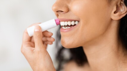 Naklejka premium Smiling Young Black Woman Applying Pink Lip Balm With One Hand While Holding A Tube In Other Hand At Home, In A Casual Beauty Setting, Engaging In Self-care Routine. Beauty Concept.