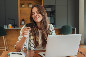Naklejka na ściany i meble Young woman smiling, holding a credit card and using a laptop computer for online shopping, confirming a purchase on an e-commerce website at home