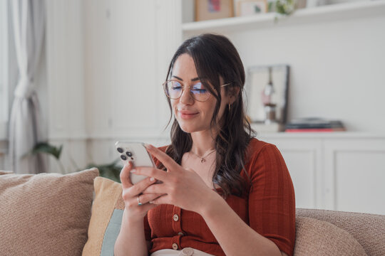 A young woman relaxes on a couch in a bright living room, smiling as she scrolls her smartphone and messages friends, enjoying a cozy, casual moment of digital connection and leisure.