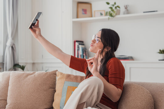 Young woman sitting on a comfortable sofa at home, holding a smartphone to capture a selfie, expressing herself with a peace sign gesture and puckered lips for social media
