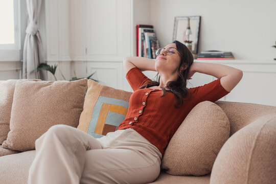 A young woman lounges on a cozy sofa with eyes closed and hands behind her head, enjoying a calm, comfortable moment of relaxation and quiet leisure in a bright, modern living room.