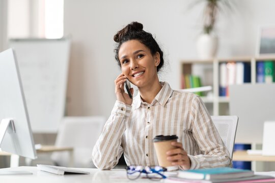 Smiling African American Woman Engaging In Phone Conversation While Holding Coffee Cup At Her Desk In Modern Office Environment, Feeling Happy And Focused, Business Concept.
