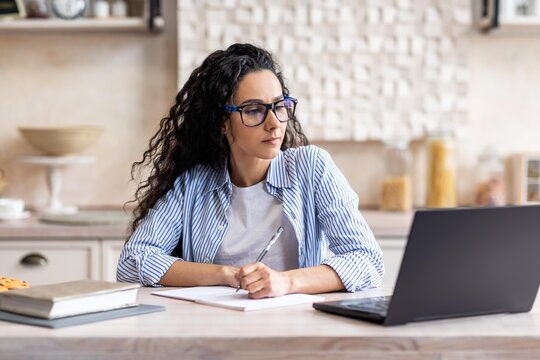 Focused Hispanic Woman In Her Early 30s Taking Notes While Working On Laptop At Home Office. Expressing Concentration And Engagement In Task At Modern Workspace. Business Concept.