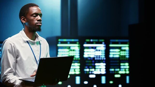 African American computer scientist walks among server cabinets in a high tech facility, overseeing computing resources during computer expert patching server.
