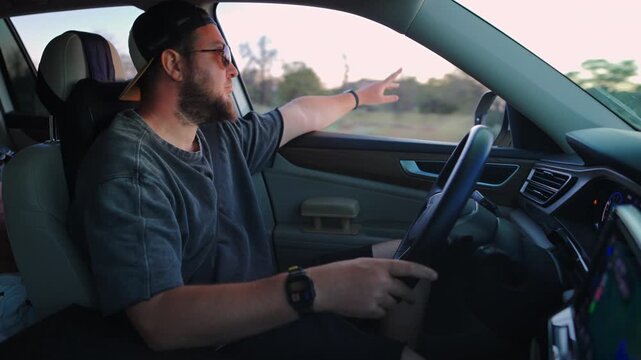 Bearded man wearing sunglasses and backwards cap drives car while pointing out passenger window. Interior view shows leather seats and dashboard during evening rural drive.