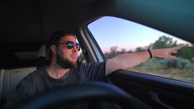 Bearded man in backwards black cap and reflective sunglasses enjoys a scenic drive through arid desert terrain at dusk, his arm surfing the warm evening breeze outside the open car window.