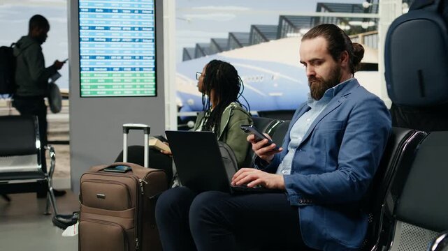 Businessman uses a laptop while waiting for a smartphone message at the airport, checking notifications and completing tasks before his departure flight as a business traveler near the gate.