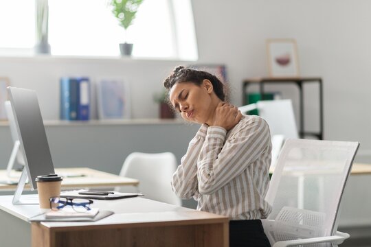 Worried Black Woman Experiencing Pain While Touching Her Neck, Sitting At Desk In Office Environment. She Shows Signs Of Discomfort And Stress, Reflecting Common Office Health Issues. Healthcare